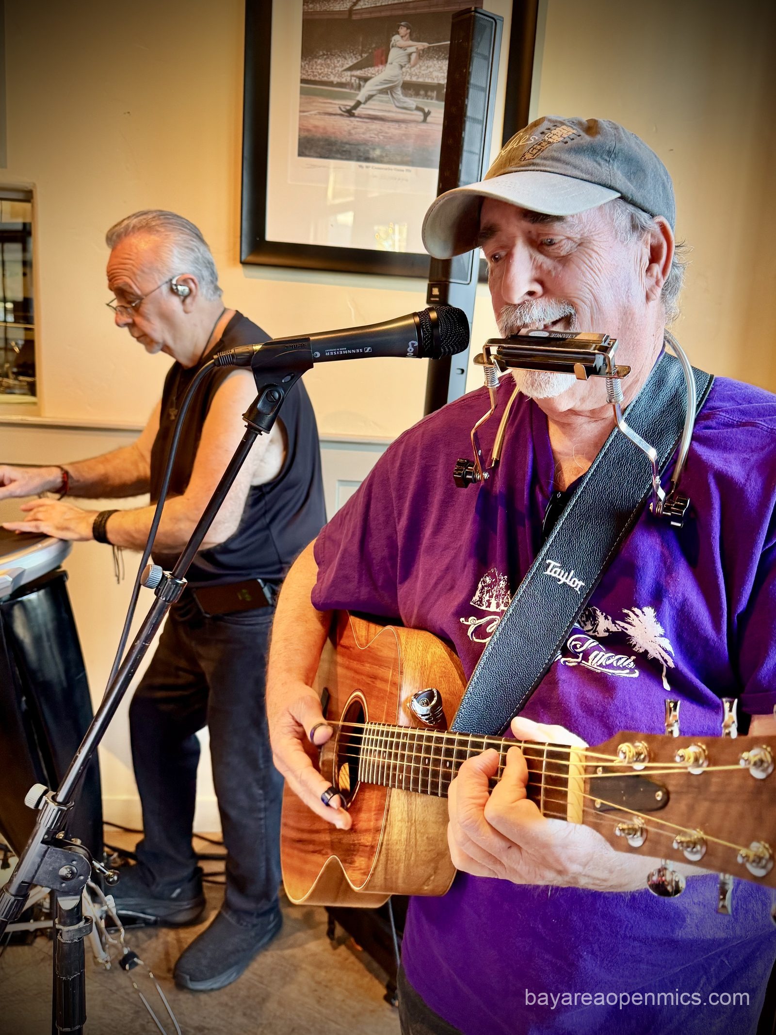 a man in a purple shirt smiles while playing guitar, in the background a man in a sleeveless black tee plays percussion