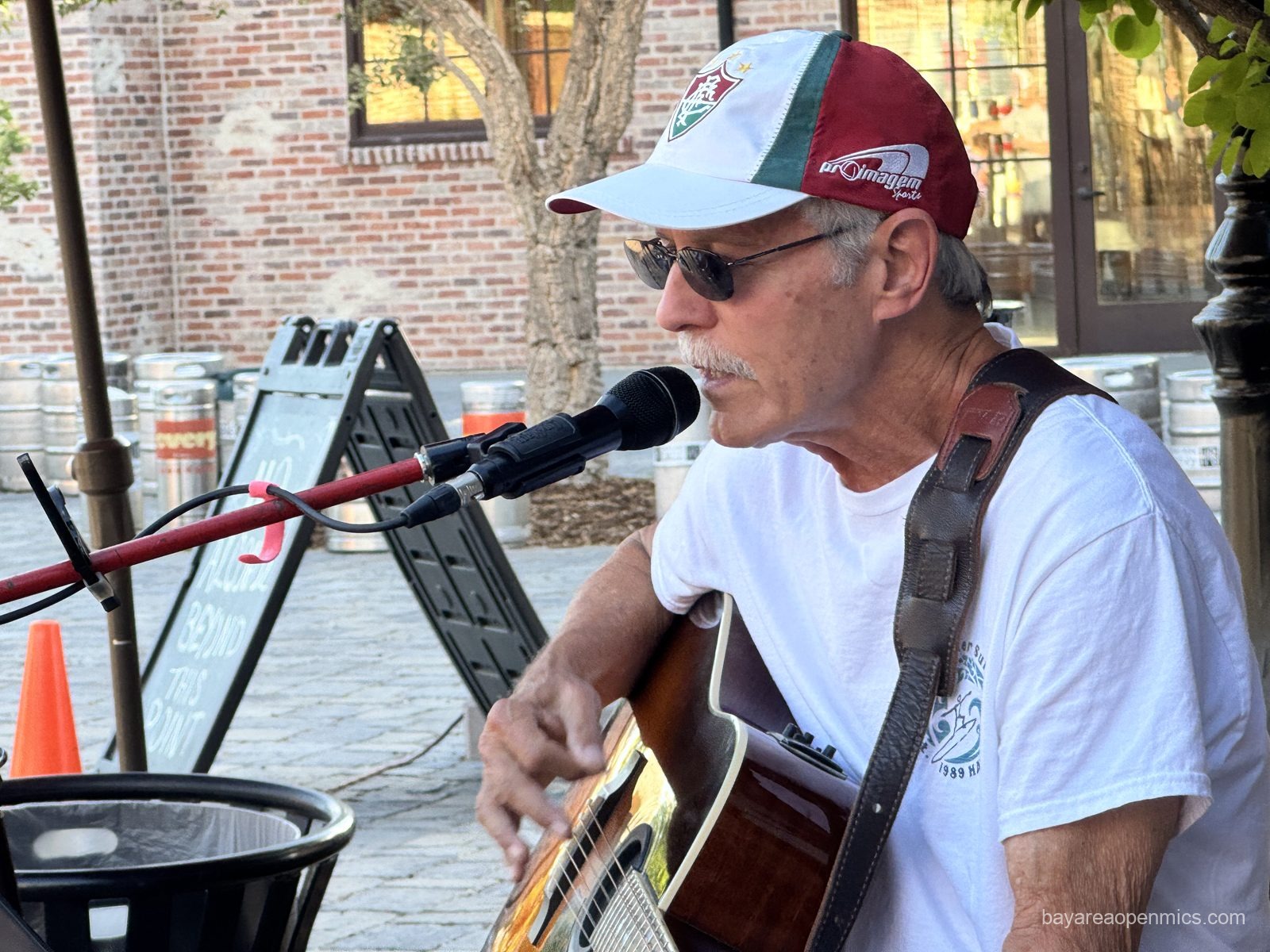 a man with a grey mustache in a baseball cap and sunglasses strums a guitar while singing into a microphone 