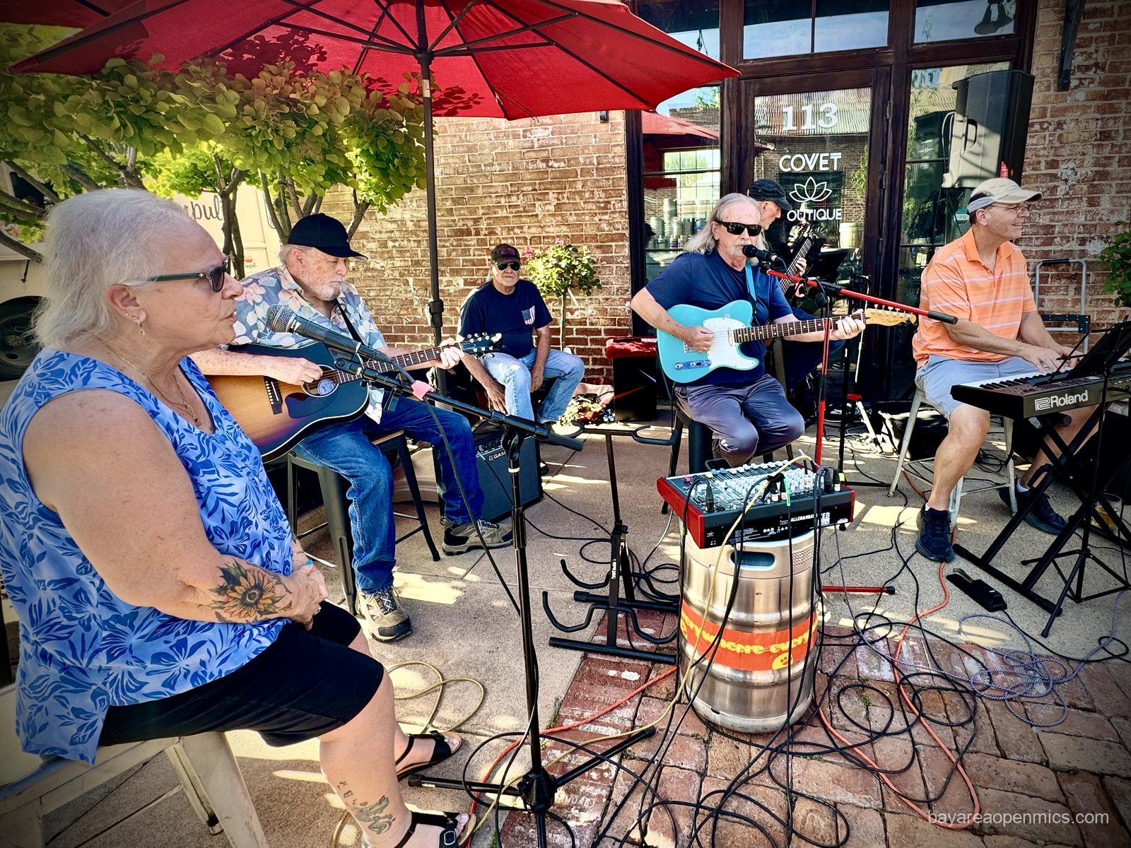 A mixing board is perched on a steel beer keg in the midst of six older rock musicians gathered around an assembly of microphones and cords under a large red sun umbrella