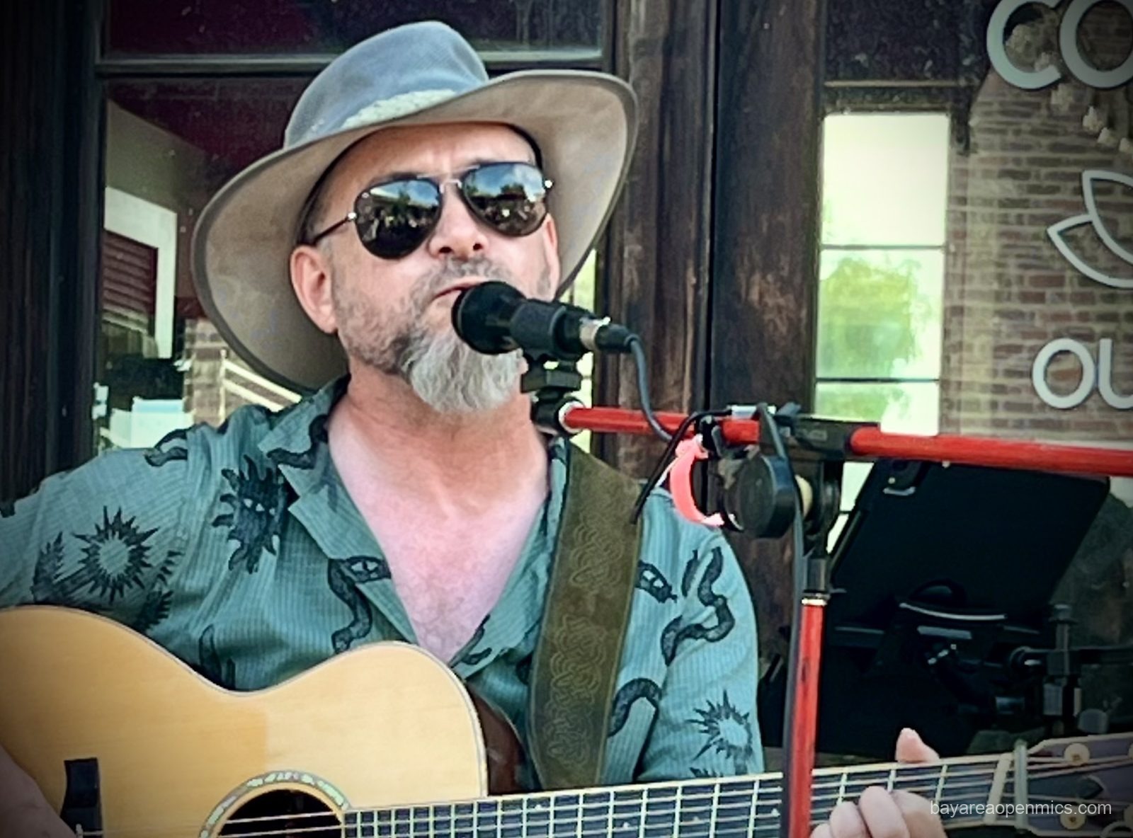 a man wearing sunglasses and broad-brimmed hat sings and plays guitar.