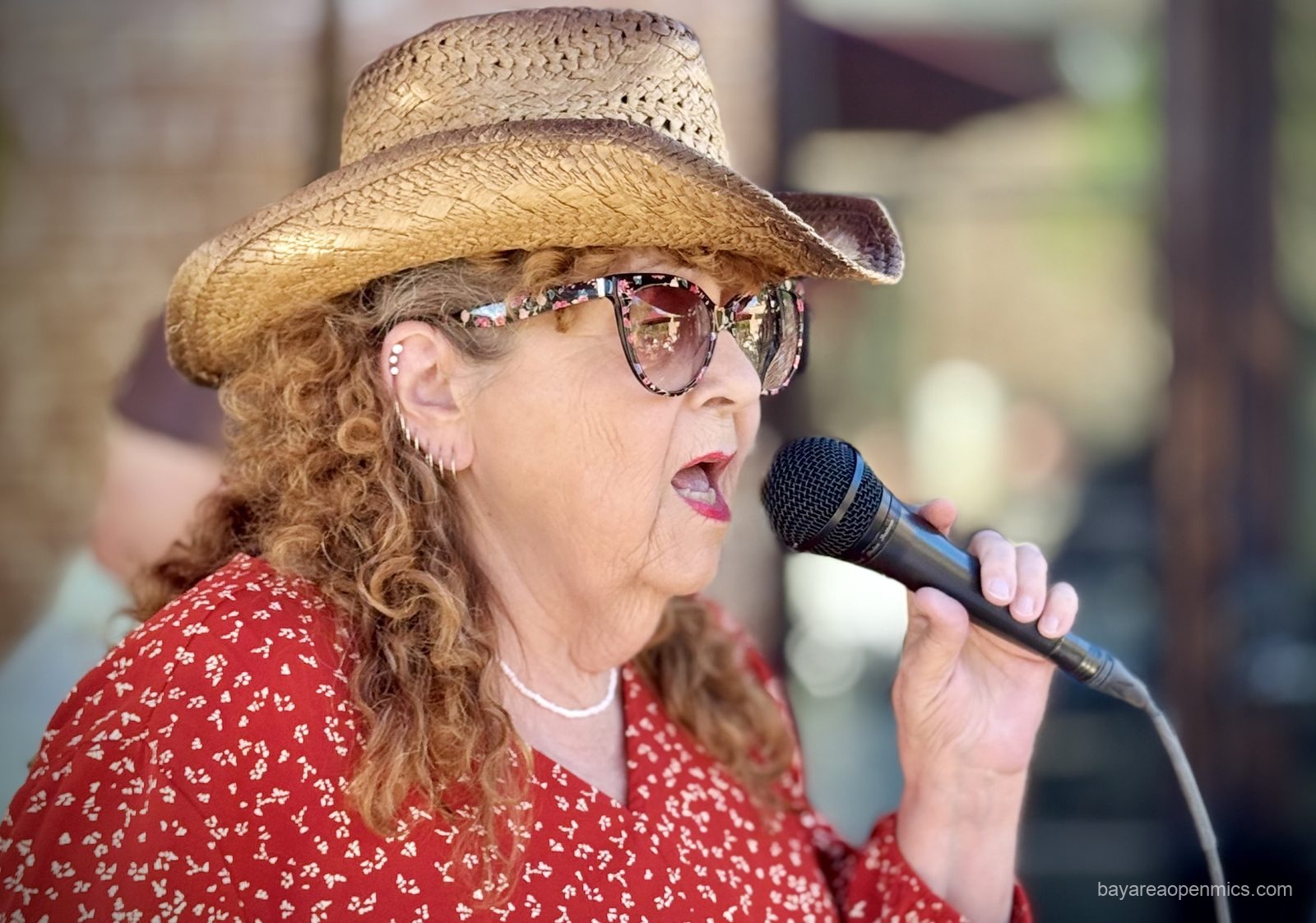 a woman in a red blouse, straw cowboy hat, and floral-print glasses hold a microphone while singing 