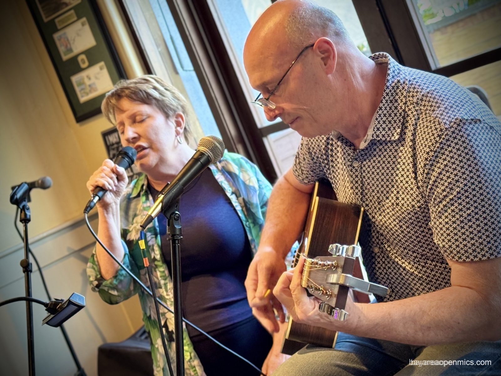 Laura holds a microphone while singing with her eyes closed, in the foreground Sean A smiles while playing guitar at Capers open mic in Campbell