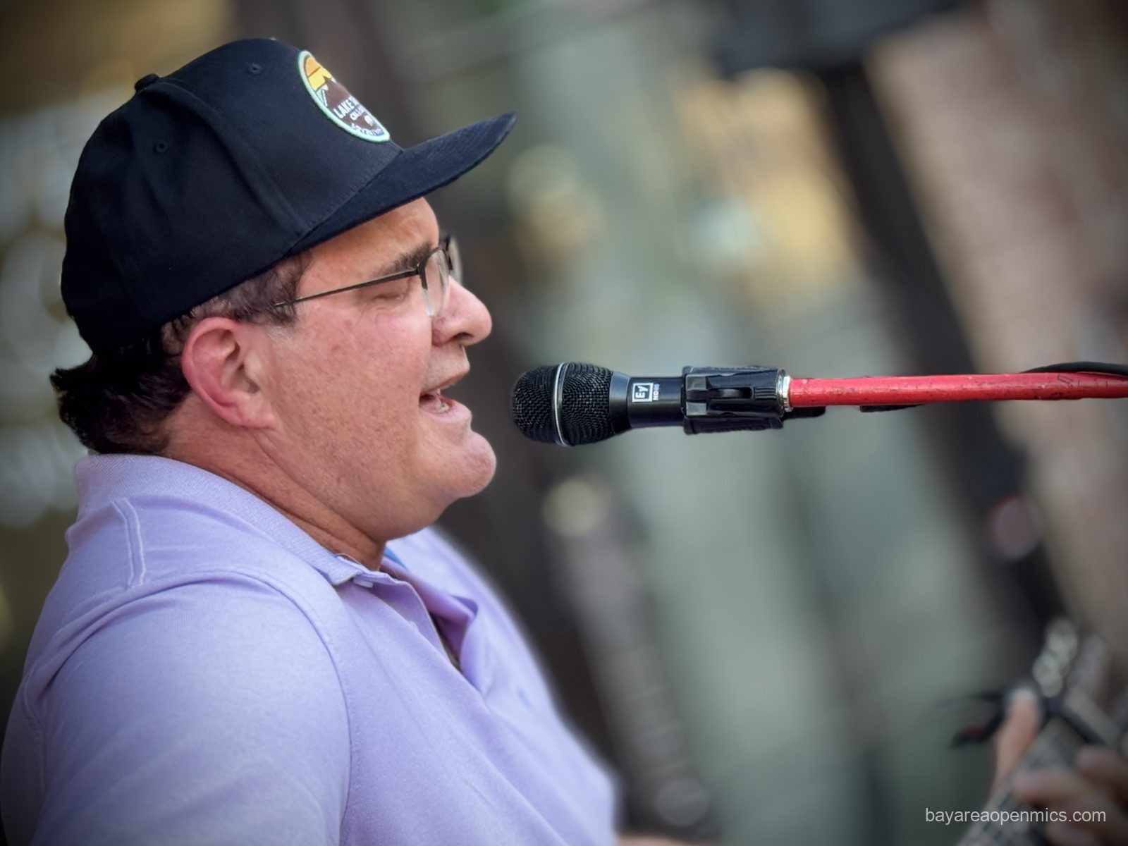 A man in a black baseball cap and glasses sings passionately into a microphone while strumming a guitar