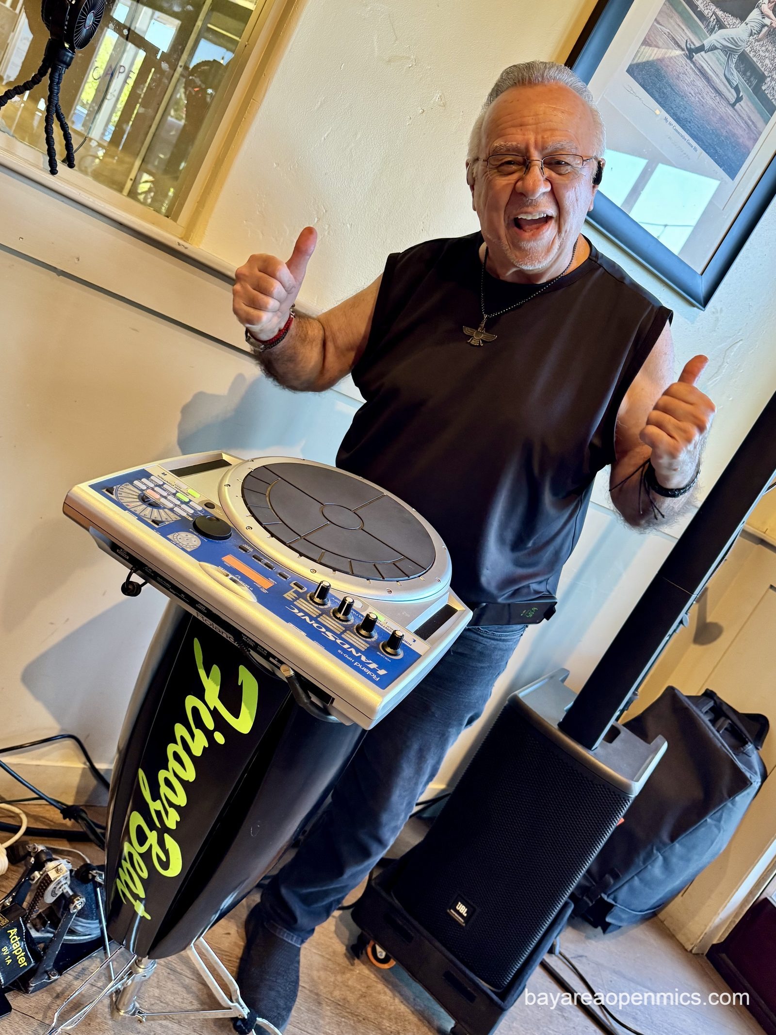 a man in black sleeveless tee and glasses with a zoroastrian medallion around his neck gives two thumbs up while standing behind a congo-shaped electronic percussion set up