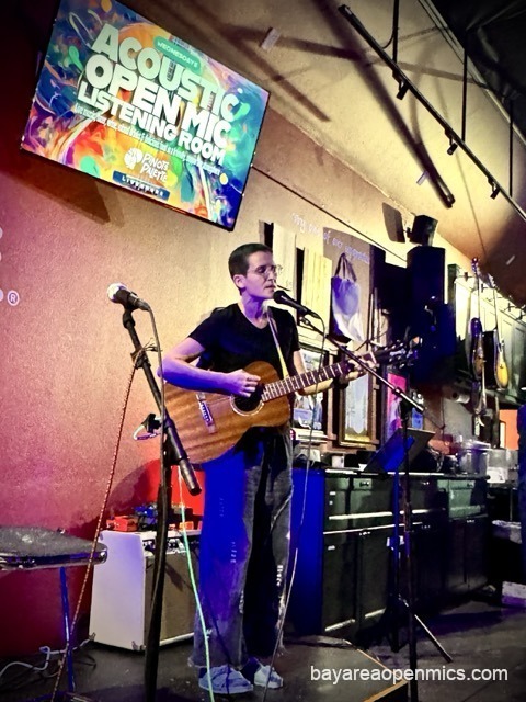 A woman with a buzz cut and baggy jeans performs onstage beneath the Acoustic Open Mic Listening Room sign at Pinot's Palette