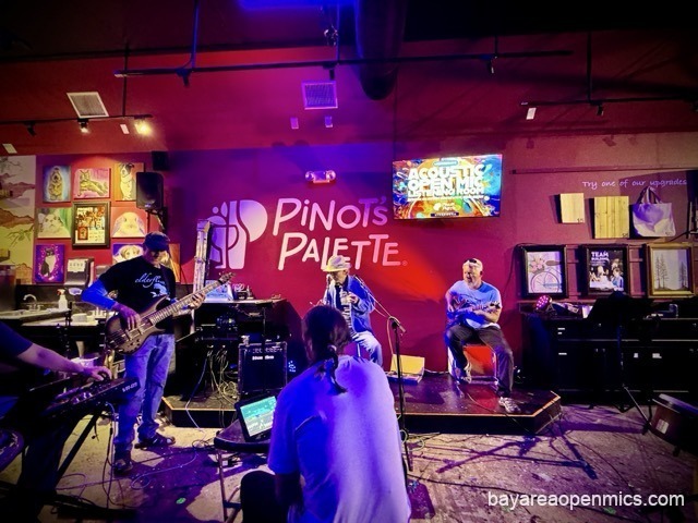 Accompanists are depicted at Pinot's Palette Open mic: an arm extends into the frame over a piano keyboard; a bass player in a baseball cap, jeans and sandals plays while standing; a lead singer in a cowboy hat holds a harmonica; an electric lead guitar player with short white hair and a pair of sunglasses on his head jams along to the singer's left onstage, and in the foreground we can see open mic host John Iacono's with his back turned to the camera, a glowing iPad with his sound controls in front of him, and he is slightly bent forward, sitting atop te cajon he is playing