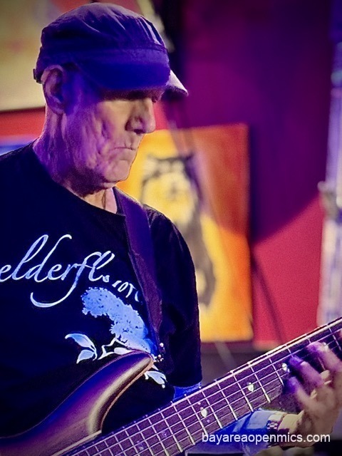 An older man in a snug cloth baseball cap plays a six-string bass guitar while wearing a shirt that reads, 'Elderflower.' at Pinot's Palette open mic in Livermore