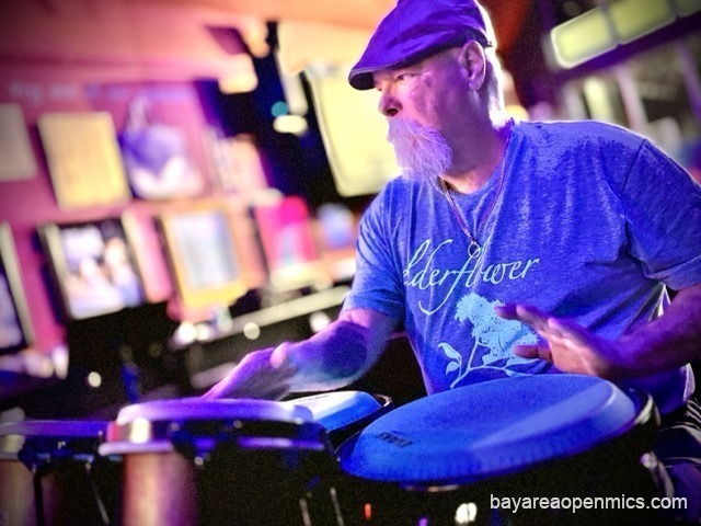 A bearded man in a flat cap expertly plays congas with bongos in the foreground at Pinot's Palette