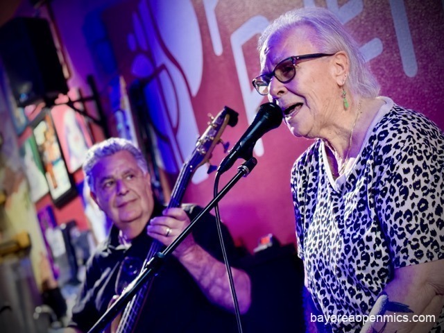 An older woman in a leopard-print top sings into a microphone while a white-haired bassist accompanies her, smiling, in the background