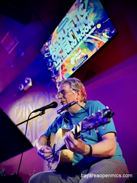 Richard Lappin with a harmonica bracket around his neck sings while playing acoustic guitar beneath the Acoustic Open Mic Listening Room sign
