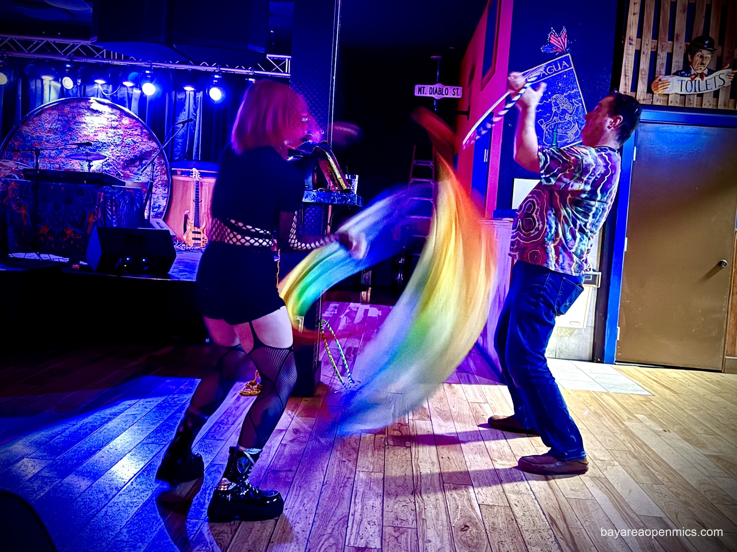 An image of a woman in a black outfit with fishnet tights and shiny military boots facing away from the camera spins colorful rainbow flags, which blur with motion, while a nearby man leans back while twirling a hula hoop with both bands