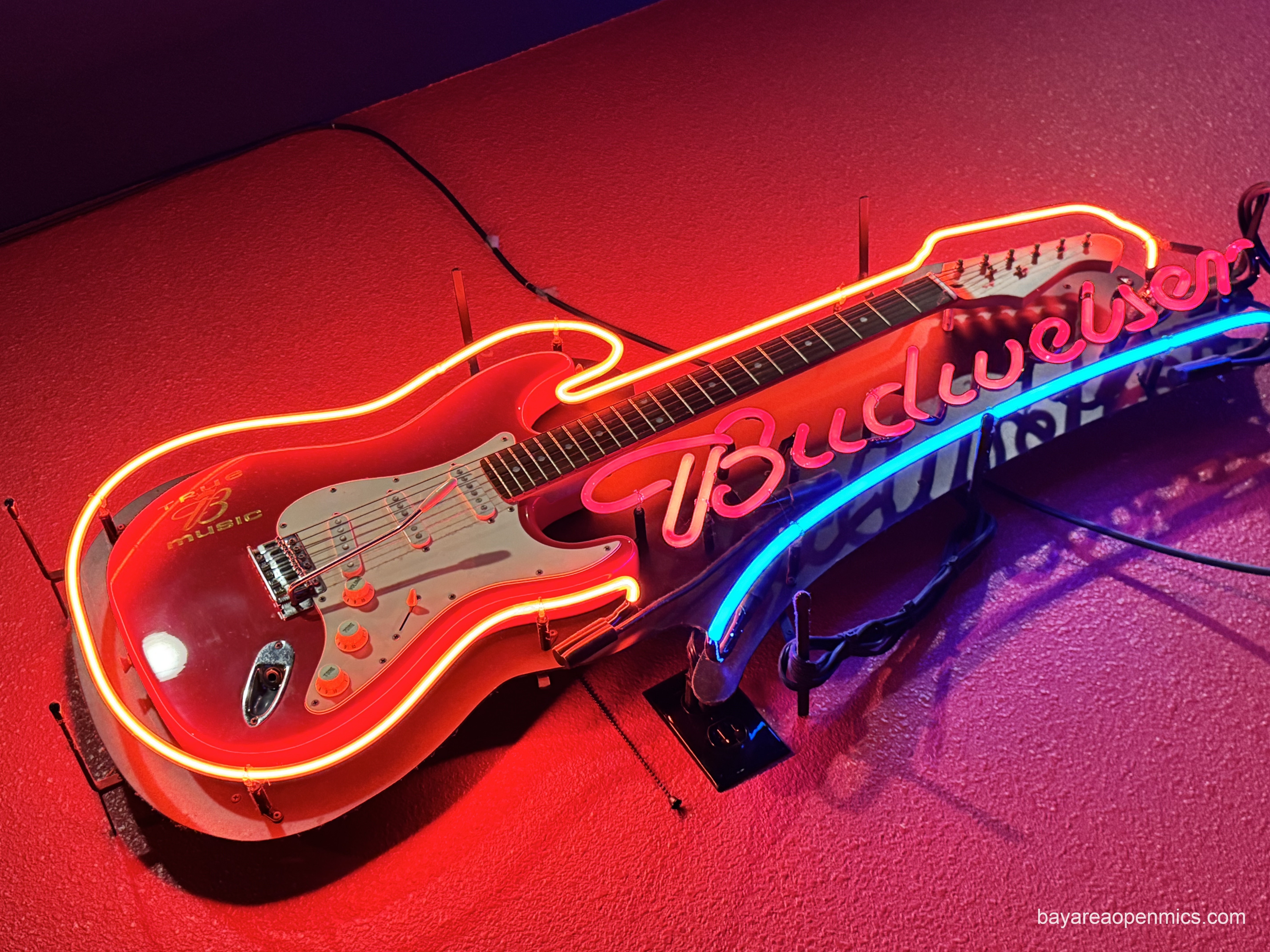 A red stratocaster with a whammy bar and a 'B MUSIC' logo on the body is mounted on a backing board and outlined in red neon next to a cursive version of the word 'Budweiser,' also in red neon and underlined by a blue neon swoosh. You can see the power cords extending away from the sign up to the ceiling