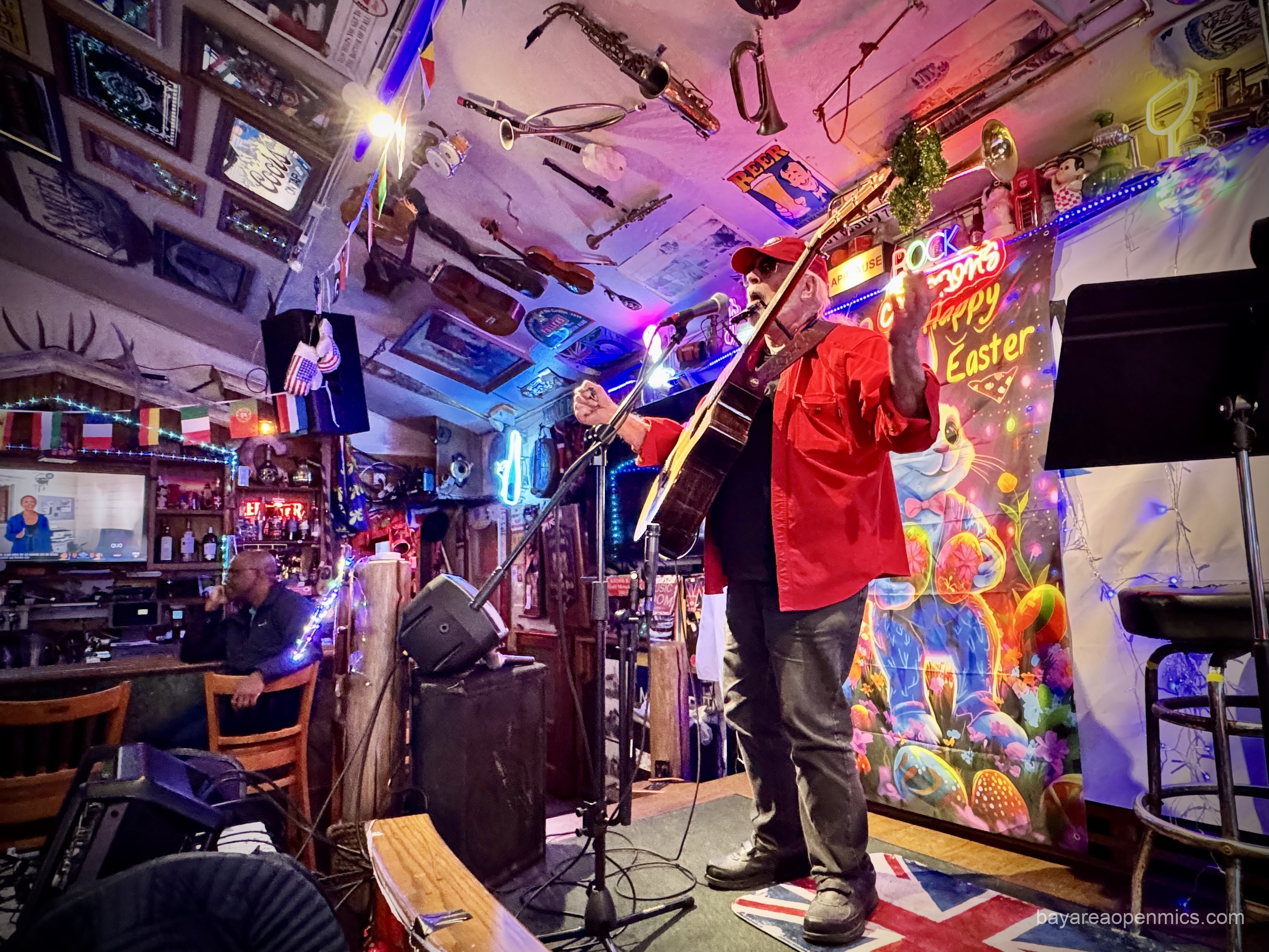 An older man wearing a SF Giants baseball cap and red jacket with a harmonica holder around his neck and a guitar around his shoulders throws his arms wide onstage