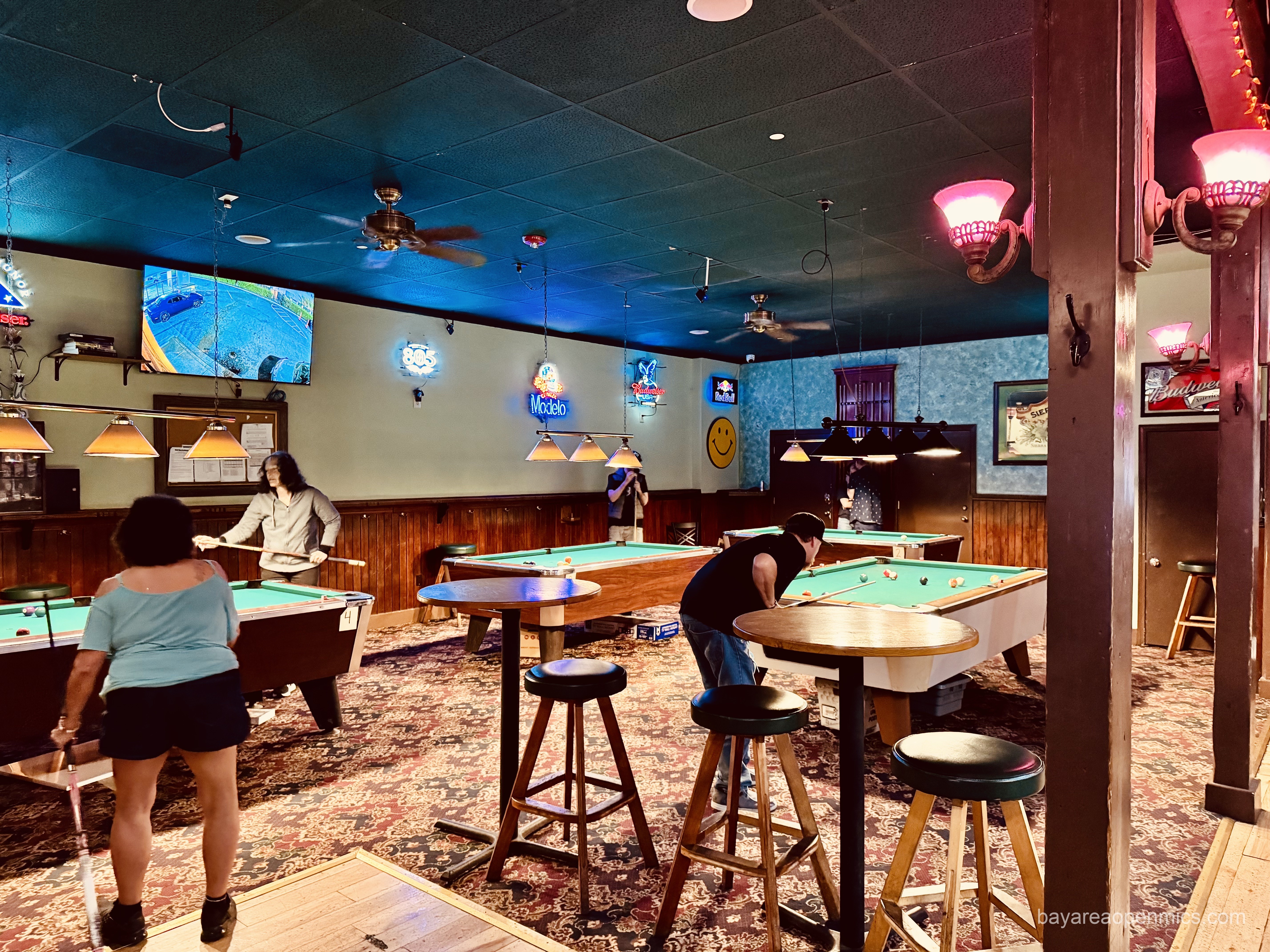 Four pool tables and folks playing pool are depicted in a carpeted room with high top tables and stools